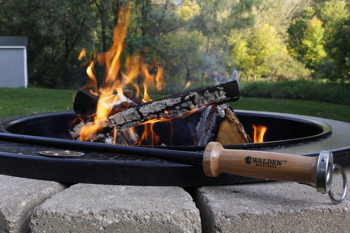 A Walden-branded Stoker Poker resting across the rim of a lit fire pit, flames climbing off split logs